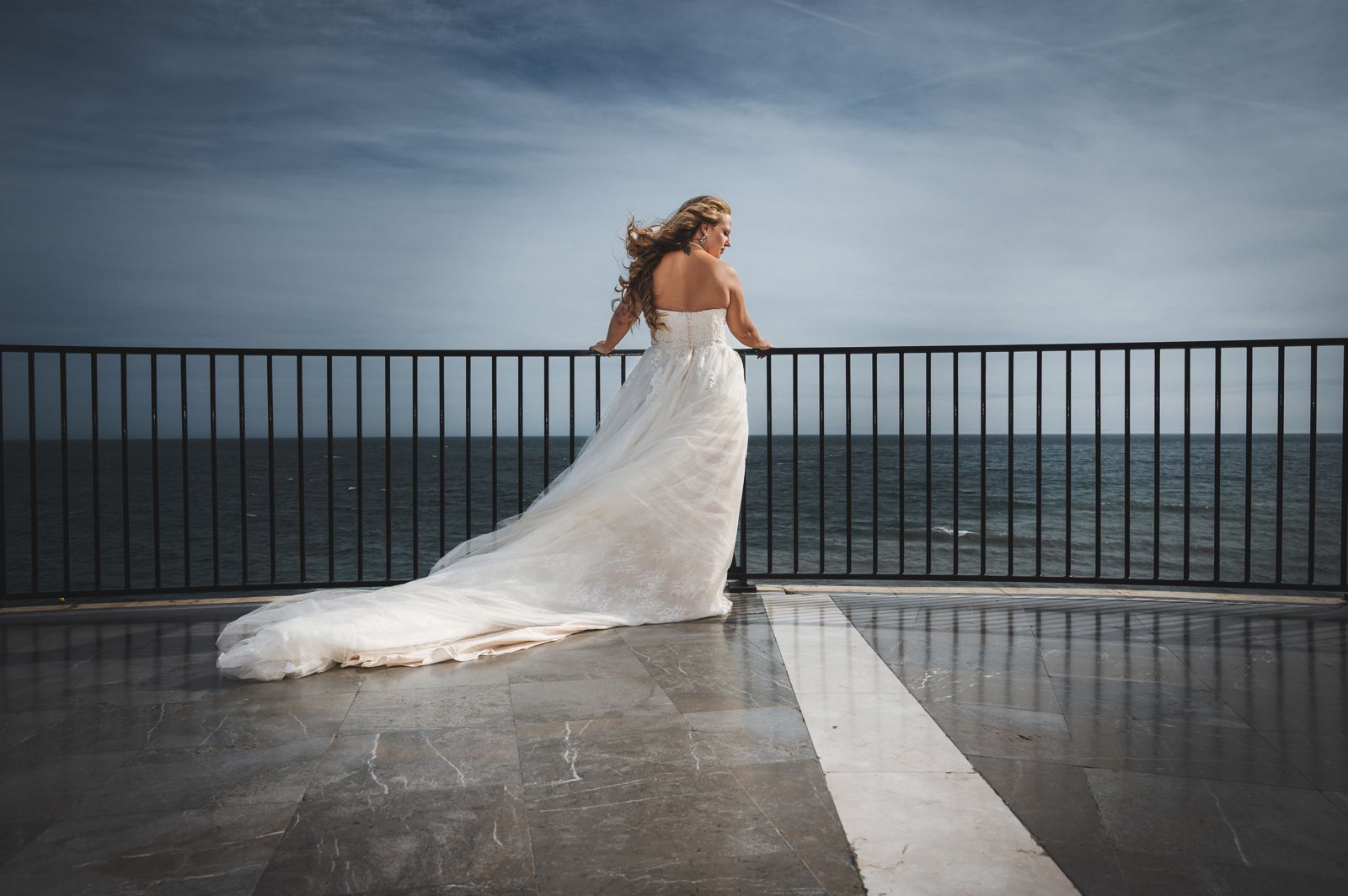 La novia, de espaldas, se apoya en la barandilla de un mirador frente al mar con la cola del vestido extendida