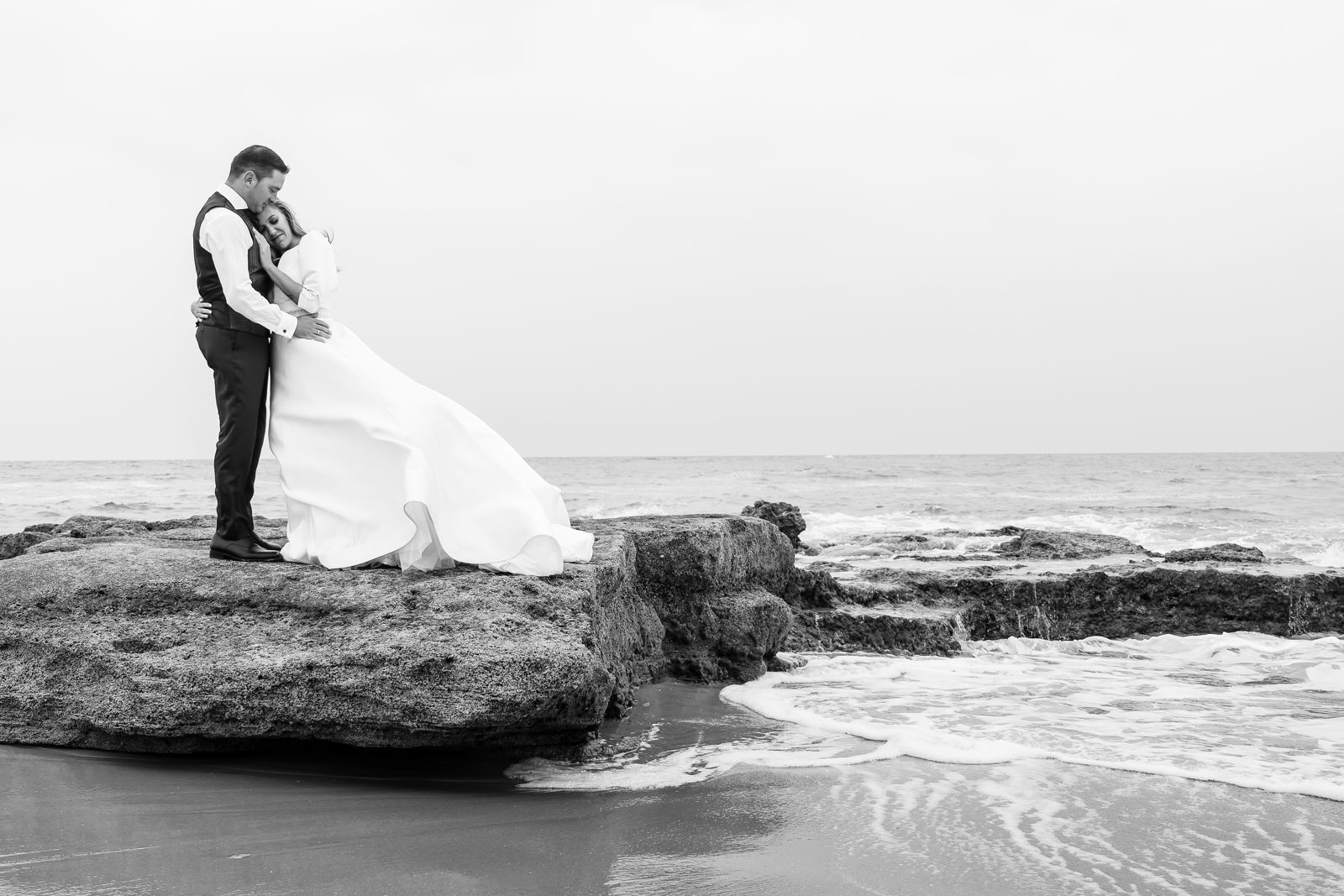 Pareja abrazada sobre una roca junto al mar, el vestido movido por el viento