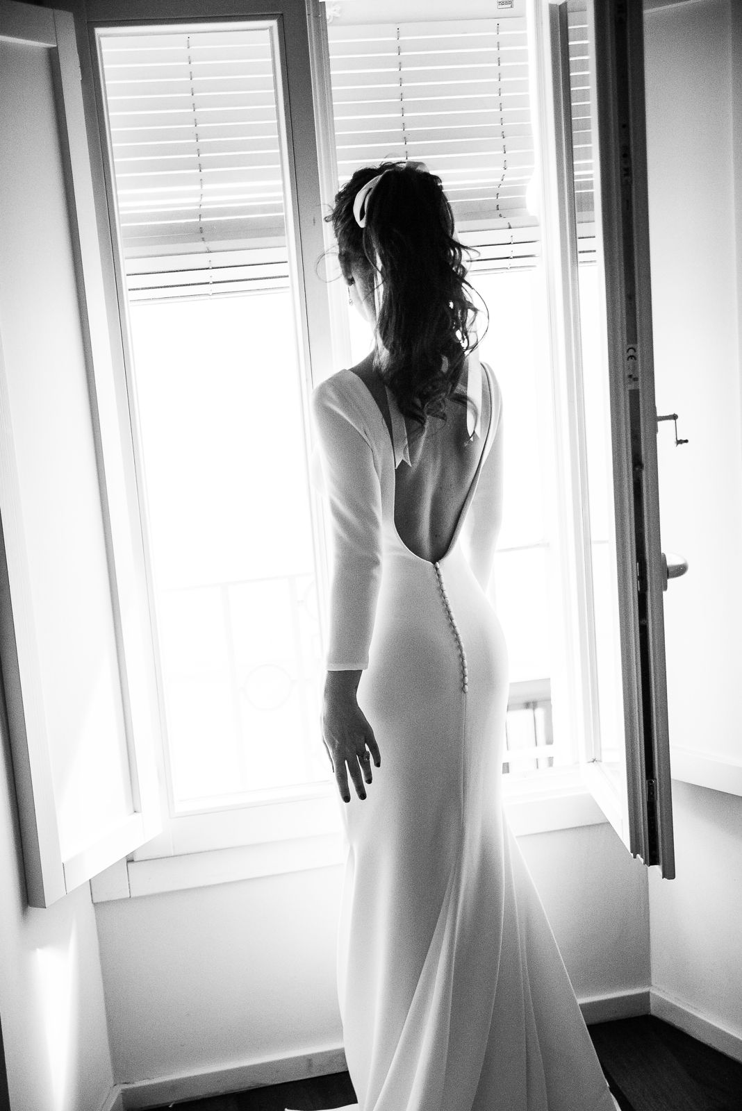 La novia, de espaldas, mira por una ventana abierta a contraluz con el vestido de espalda descubierta